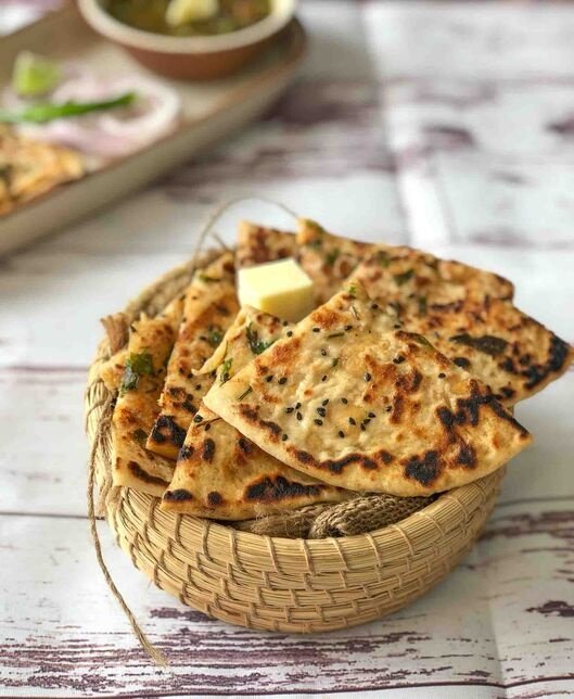 A woven basket filled with pieces of freshly baked and buttered Indian flatbread garnished with herbs and black sesame seeds.