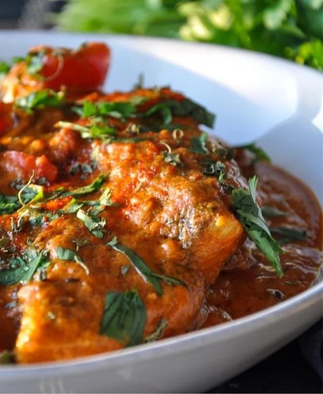 A white bowl containing tender salmon fish cooked in a savory, spiced tomato curry sauce and garnished with fresh herbs.